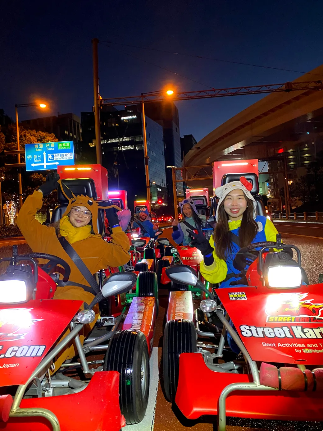 Group of people in colorful costumes posing on red street-kart vehicles at night in a city street parade.