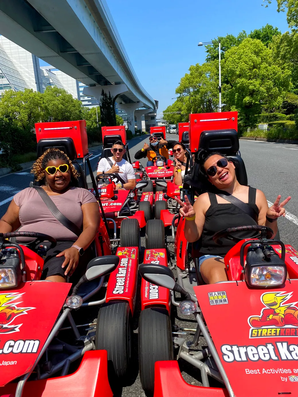 Group of six friends in bright red go-karts on a city street, smiling at the camera under a raised transit track as they pose for a fun ride.