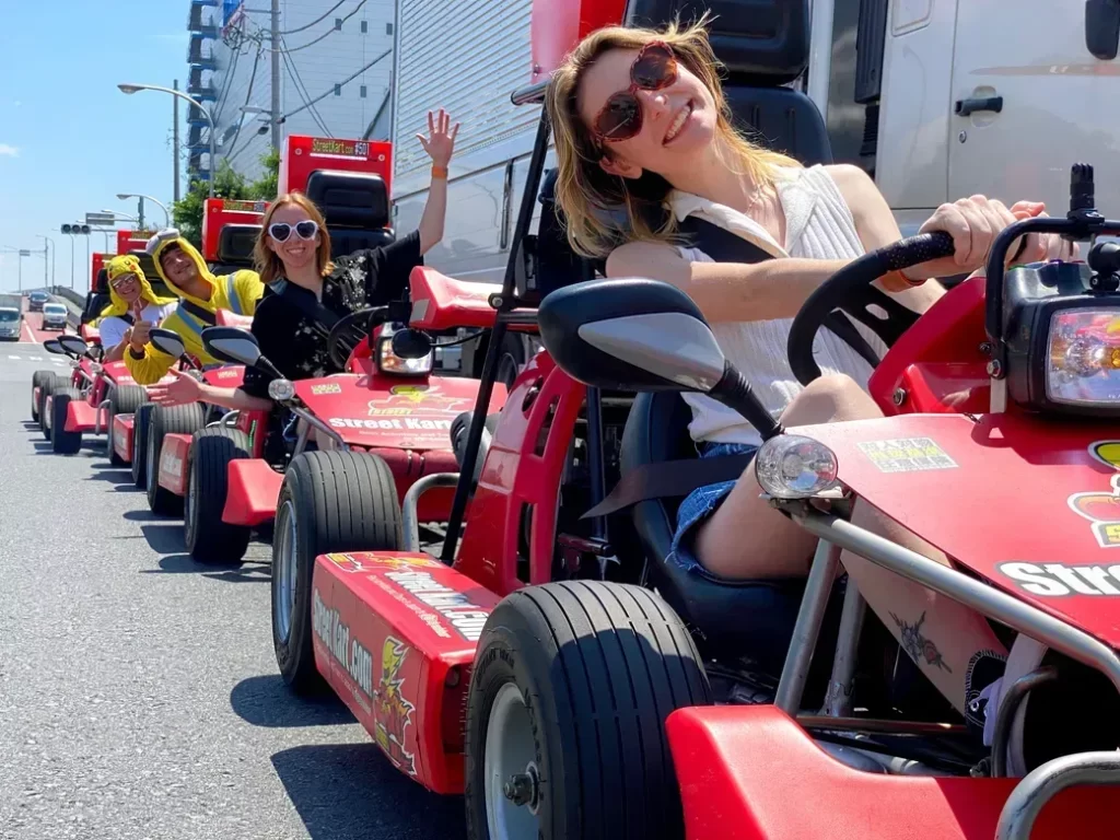 IMG_5020 - StreetKart A group of people in red go-karts driving in a sunny city parade, the woman in the front smiling and waving from her kart.